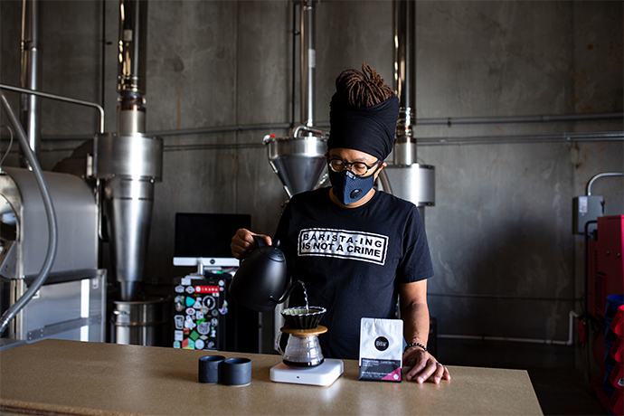 Barista pouring water from a goose-necked kettle into a V60 pourover brewer with Black & White coffee beans next to it.