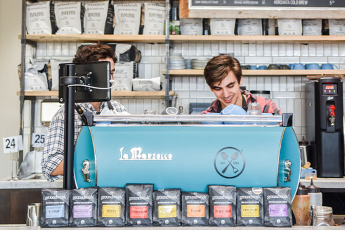 Two baristas standing behind a blue La Marzocco espresso machine with Groundwork coffee bags in foreground.