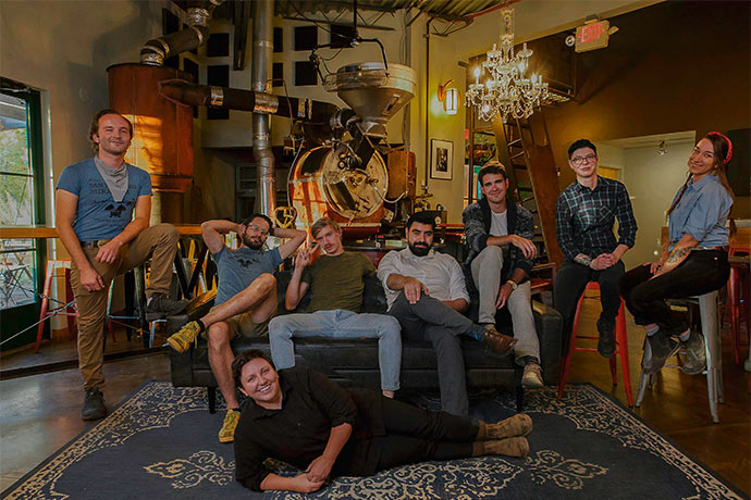 Iconik Coffee Roasters team standing and sitting in various positions in front of their roasting machine.