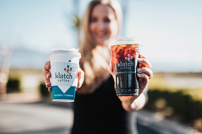 Person holding a Klatch coffee cup in one hand and a Klatch plastic cup in another.